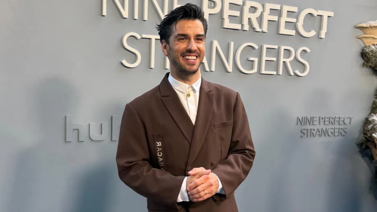 Aras Aydın, 'Nine Perfect Strangers' yazılı promosyon paneli önünde resmi pozunda. Kahverengi takım elbise ve beyaz gömlek giymiş, hafif gülümseyerek kameraya bakıyor. Ellerini önünde kavuşturmuş halde duruyor. Muhtemelen dizinin tanıtım etkinliğinden bir kare.