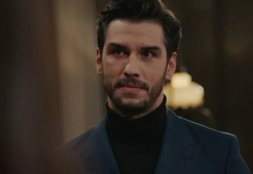 Koyu renk balıkçı yaka giymiş Aras Aydın'ın yakın plan portresi. Siyah sakalları ve dikkatli bakışlarıyla objektife bakıyor. Koyu renk bir ceket giymiş ve ciddi bir ifadeye sahip. Arka plan bulanık ve karanlık tonlarda.