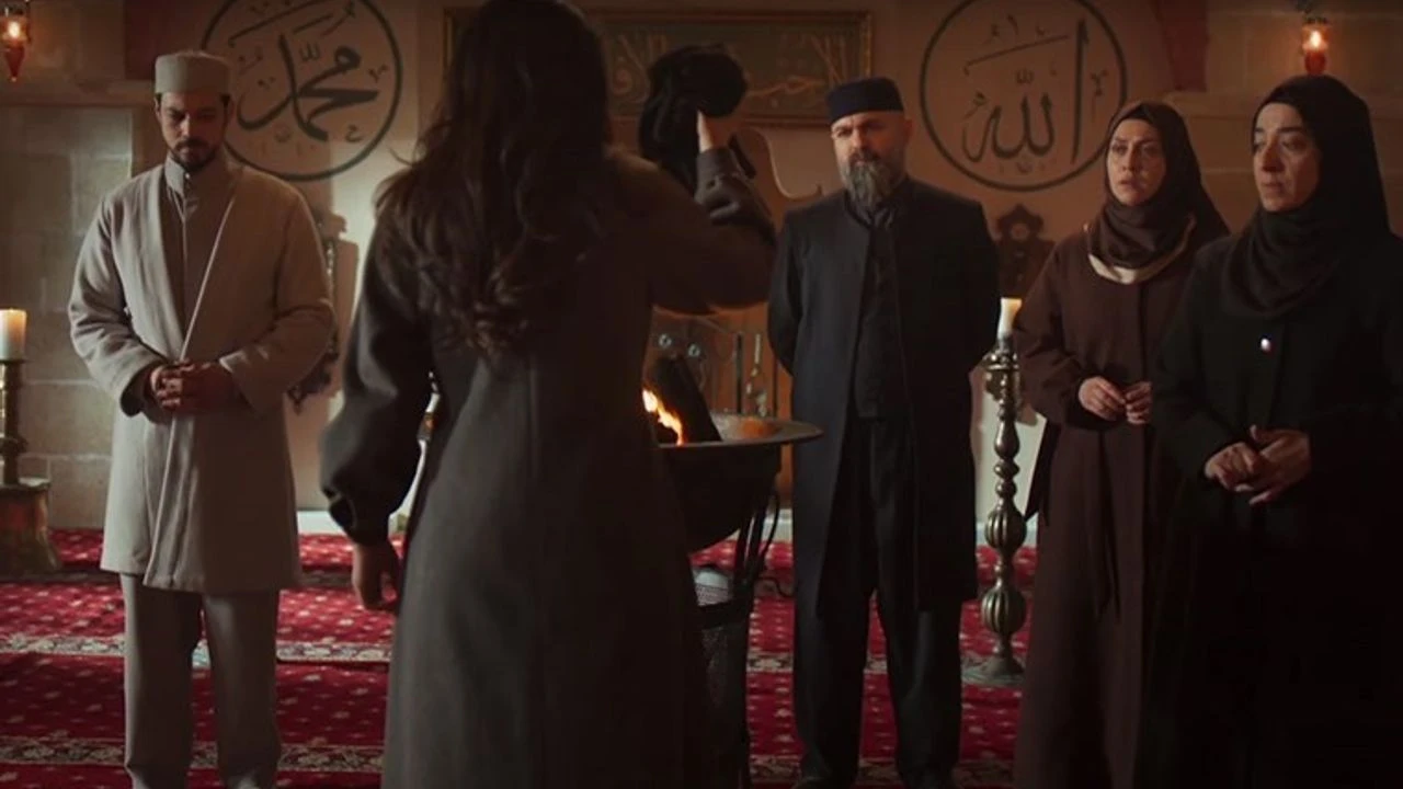 Kadın karakter, dini sembollerle süslenmiş bir mekanda elinde bir nesne tutarak karşısındaki grup ile yüzleşiyor. Arka planda İslam yazıları ve dekoratif objeler yer alıyor.