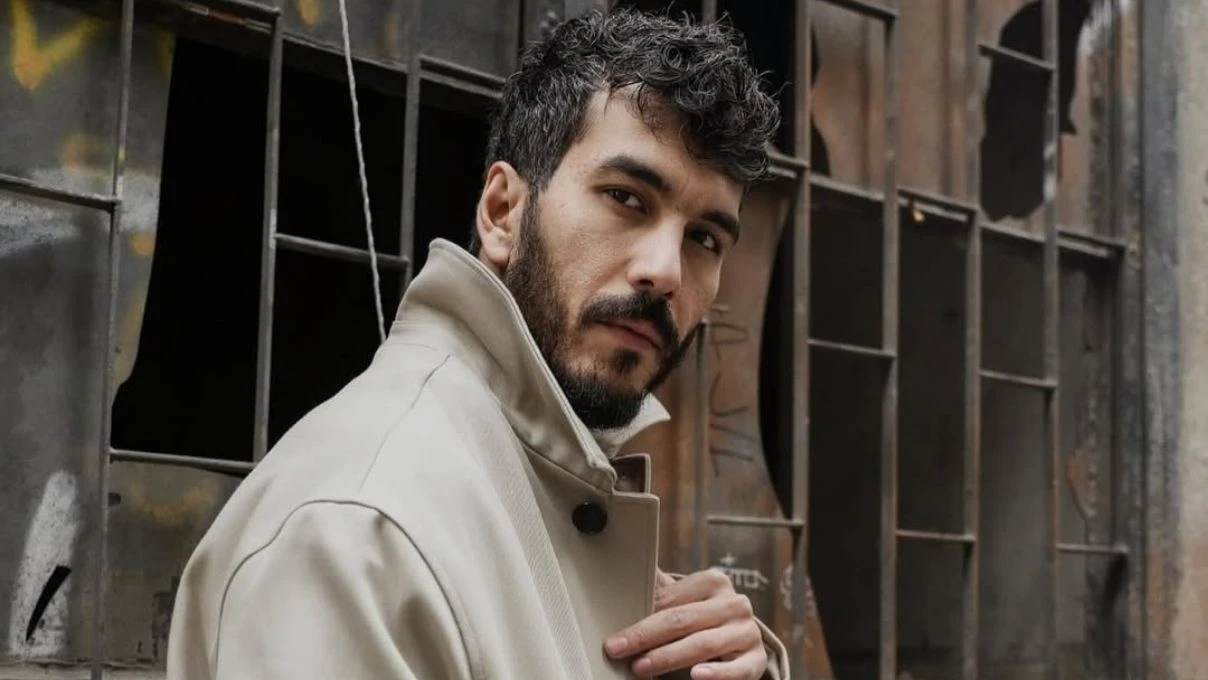 Mert Doğan oyuncu fotoğrafı — bej palto ile endüstriyel arka planda